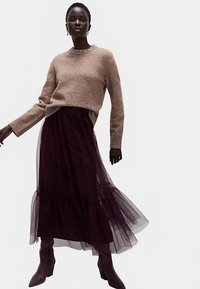 Brun strikket trøje og mørk bordeaux tulle nederdel med lagdelte flæser. Modellen har knæhøje lilla støvler på. Enkel sølvhalskæde.