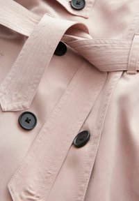 Ljusrosa trenchcoat med svarta knappar, midja med bälte och textil med synliga sömnadsdetaljer.