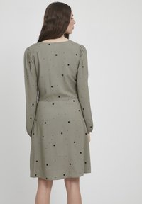 Olivengrünes Kleid mit langen Ärmeln, verziert mit schwarzen Punkten. Weicher Stoff, lockere Passform und geraffte Schulterdetails.