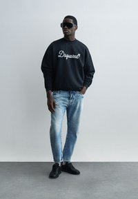 Muž v čiernej mikine Dsquared2, svetlomodrých džínsoch, čiernych teniskách a veľkých čiernych slnečných okuliaroch na jednoduchom sivom pozadí.