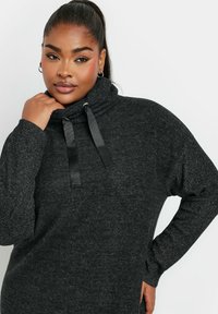 Maglione sovradimensionato grigio scuro con colletto alto, caratterizzato da lacci in satin neri e maniche testurizzate. Tessuto morbido con una vestibilità rilassata.