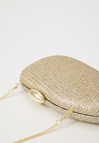 Clutch tejido de oro metálico con una textura suave, cierre ovalado y correa de cadena dorada. Forma rectangular redondeada.