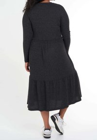 Robe longue à manches longues gris foncé à nervures avec une jupe évasée, présentant une taille cintrée et un bas à volants. Portée avec des baskets blanches.