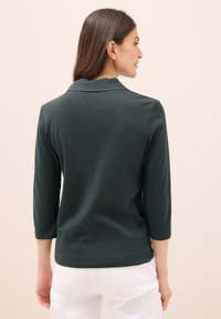 Tmavě zelené polo tričko s vlnitým límcem a tříčtvrtečními rukávy, vyrobené z jemné látky. Má hladký povrch a přiléhavý střih.
