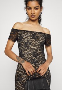 Vestido preto de renda com decote ombro a ombro, adornado com um padrão floral sobre um tecido bege. A modelo segura uma bolsa preta, destacando a intrincada textura da renda.