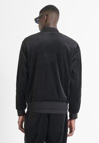 Chaqueta bomber de velour negra con cuello alto, puños y dobladillo acanalados, con un estampado tonal "CLUBBER" en la espalda. Textura suave.