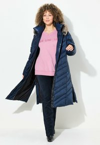Marinblå lång pufferjacka med dragkedjestängning och lurvigt foder i huvan, i kombination med en rosa V-ringad sweatshirt och svarta manchesterbyxor.