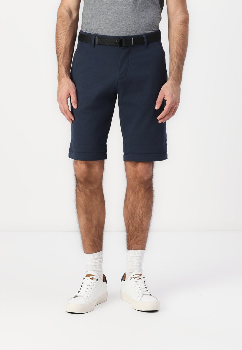 Shorts bleu marine en tissu doux, avec des ourlets roulés, une ceinture noire, portés avec des baskets blanches et des chaussettes.