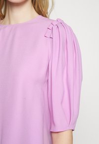 Blusa lilla chiaro con maniche corte a sbuffo. Presenta un tessuto morbido, scollo rotondo e dettagli plissettati sulla spalla.
