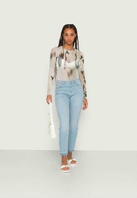 Camisola de manga comprida em bege com padrões abstratos em cinza e castanho, combinada com jeans skinny azul claro e sandálias brancas de salto bloco.