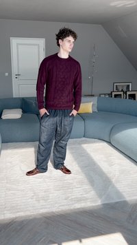 Jersey de punto burdeos oscuro con un patrón texturizado, pantalones grises de corte holgado y zapatos marrones, de pie sobre una alfombra clara en un interior moderno.