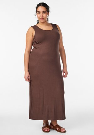Kobieta o kręconych włosach, ubrana w bezrękawnikową brązową sukienkę maxi i brązowe sandały z krzyżującymi się paskami, stoi na jednolitym białym tle.
