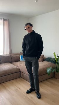 Jeune homme aux cheveux noirs et bouclés, portant un pull zippé noir, un pantalon gris et des chaussures noires, debout à l'intérieur près d'un canapé marron et d'une plante.