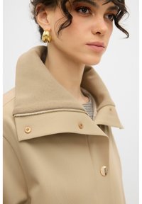 Beige trenchcoat met een grote, gevouwen kraag, gouden hardware en een ritssluiting aan de voorkant. De stof lijkt glad met een gestructureerd ontwerp.