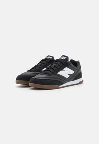 Pari di sneakers nere New Balance con logo bianco e suole marroni, allacciate, posizionate affiancate su uno sfondo bianco.