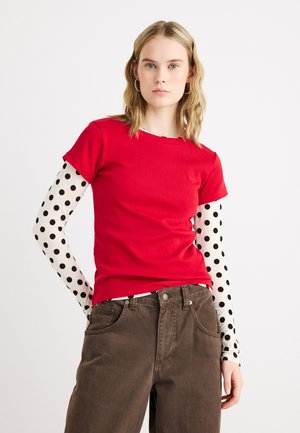 Nő piros rövid ujjú felsőt visel fehér hosszú ujjú, fekete pöttyös ingre és barna nadrágot, fehér háttér előtt áll.