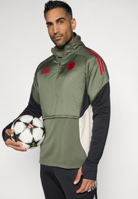 Αθλητική ζακέτα μακριών μανικιών με μισό φερμουάρ σε ελιά, με κόκκινες λεπτομέρειες και λογότυπο, σε συνδυασμό με μια μαύρη και λευκή μπάλα ποδοσφαίρου.