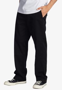 Black fabric trousers with a loose fit, two side pockets, and a smooth texture, paired with black sneakers featuring white laces.