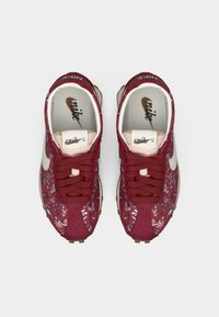 Crvene Nike tenisice s uzorkom, s gornjim dijelom od teksturirane tkanine, bijelim swoosh logotipom i smešnim gumenim potplatom. Imaju okrugle vezice i podstavljeni jezik.