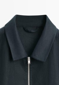 Col de veste bleu marine foncé avec une fermeture éclair métallique argentée partiellement fermée, montrant un tissu texturé et des détails de couture soignés.