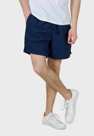 Mand iført marineblå shorts med snøre, hvide sneakers og en hvid t-shirt med benene krydset mod en ensfarvet baggrund.