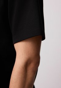 Zwarte kortekorte mouw shirt met een soepel textuur en gestikte naden, gepositioneerd naast een lichtgekleurde arm.