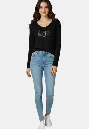 Giovane donna con maglia nera a maniche lunghe con logo "Liu Jo" e skinny jeans azzurri chiari, in piedi e sorridente, sneakers bianche visibili.