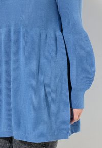 Blauer gestrickter Pullover mit einem ausgestellten Peplum-Design, gerippter Textur und langen Ärmeln. Seitenschlitze sichtbar, kombiniert mit grauen Jeans.