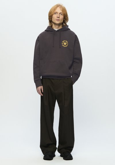 Jeune personne avec des cheveux roux mi-longs portant un sweat à capuche noir avec un logo jaune, un pantalon noir ample et des chaussures noires se tenant contre un fond blanc.