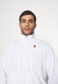 Hombre con cabello corto y oscuro y una ligera barba que lleva una chaqueta blanca con cremallera y un pequeño logo cuadrado colorido en el pecho.
