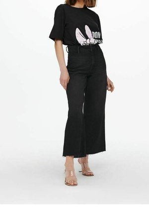 Vrouw draagt zwarte wijde jeans met hoge taille, zwart T-shirt met opdruk en doorzichtige open-teen hakken, staand tegen een witte achtergrond.