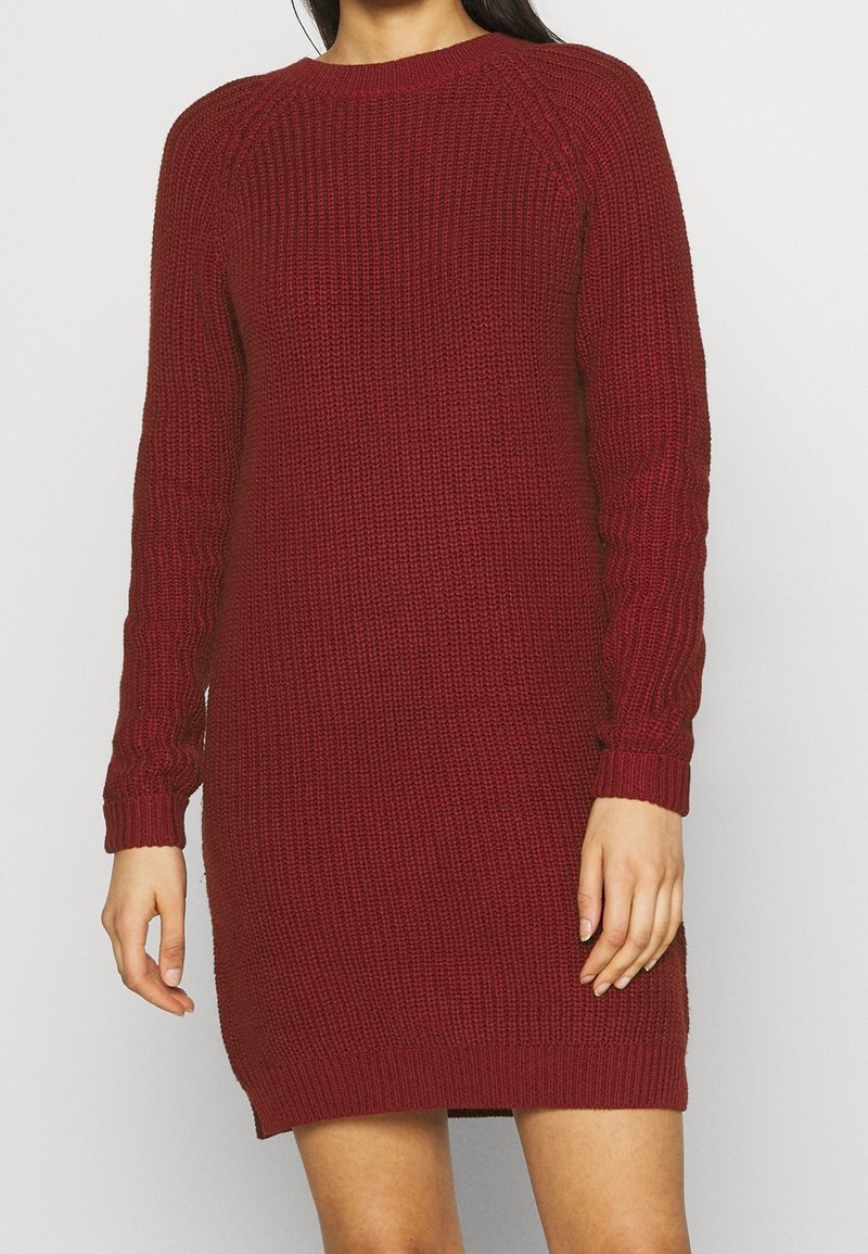 Femme portant une robe-pull en maille bordeaux à manches longues avec un col rond, debout devant un fond clair uni.