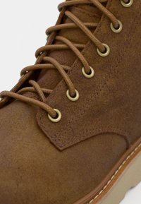 Bottine en cuir marron avec une surface texturée, dotée d'œillets en laiton, de lacets marron et d'une semelle en caoutchouc beige clair.