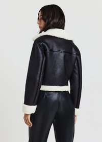 Veste noire en simili cuir coupée avec col et poignets en fausse fourrure blanche. Présente un dos sans couture et des manches larges.