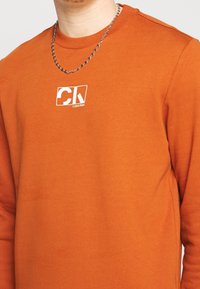 Sudadera naranja de manga larga hecha de algodón, con un logo blanco impreso y un cuello acanalado. La textura parece suave con detalles mínimos de costuras.