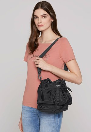 Black nylon bag with a drawstring closure, featuring a textured surface and adjustable strap. Model displays the bag over a pink shirt.