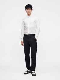 Uomo con camicia bianca abbottonata, pantaloni neri e mocassini bianchi e neri, in piedi con una mano in tasca contro uno sfondo semplice.