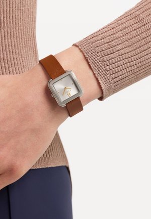 Montre carrée en argent avec un cadran blanc et des accents dorés, bracelet en cuir marron, portée sur un poignet avec un fond en pull beige à rayures.
