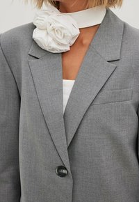 Šedý blazer z texturované látky s jedním černým knoflíkem a bílým květinovým detailem na límci.