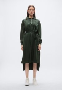 Πράσινο φόρεμα με μακριά μανίκια, τσέπες με φερμουάρ και ζώνη στη μέση. Κατασκευασμένο από λείο ύφασμα, με ψηλό γιακά και πλαϊνές σχισμές.