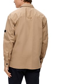 Camicia beige a maniche lunghe con colletto classico, bottoni ai polsini e una toppa nera sul braccio sinistro. Tessuto morbido e vestibilità comoda.