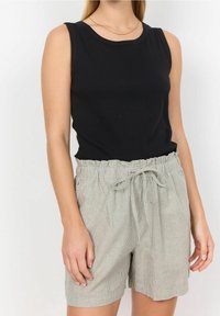 Kobieta w bezrękawniku czarnym i jasnoszarych, w poziome paski, krótkich spodniach z wysokim stanem i paskiem do związania na jednolitym tle.