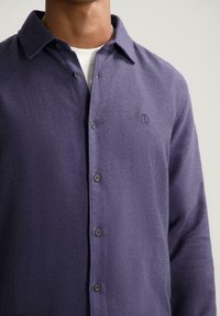 Man in een paarse button-up wolshirt over een witte crewneck, waarbij de bovenste torso en nek zichtbaar zijn tegen een effen achtergrond.