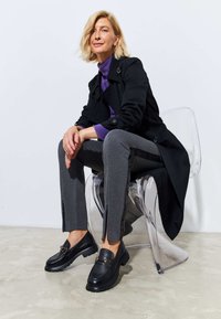 Black trench coat over purple top, gray pants with side slits, black loafers with thick soles, seated on a transparent chair.