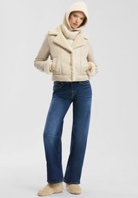 Vrouw in een beige schapenvachten jas, bijpassende met een capuchon, donkerblauwe jeans en beige pluche slippers, staand tegen een effen achtergrond.
