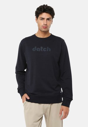 Giovane uomo che indossa una felpa nera a maniche lunghe con il logo "datch" e pantaloni beige, in piedi davanti a uno sfondo chiaro uniforme.