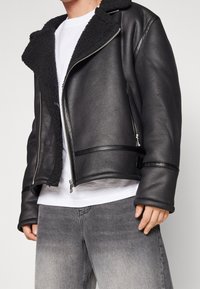 Sort læderjakke med en blød sort lammeskindskrage, lynlås foran og kontrasterende teksturerede paneler. Kombineret med falmede grå jeans.