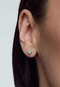 Kolczyki w kształcie serca na srebrnej podstawie, z błyszczącą powierzchnią drobnych, przezroczystych kamieni. Ujęcie z bliska na uchu.