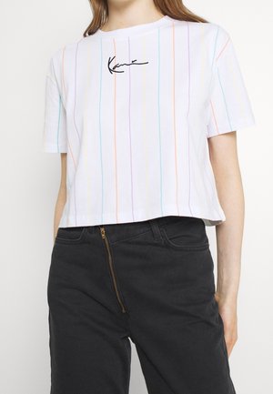 Kobieta w białym t-shircie z krótkim rękawem z cienkimi pastelowymi pionowymi paskami i czarnym logo pisanym, do tego wysokie czarne spodnie.
