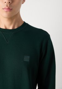Donkergroene crewneck sweatshirt van zacht materiaal, met een klein vierkant logo op de linkerkant van de borst. Eenvoudig en minimalistisch ontwerp.